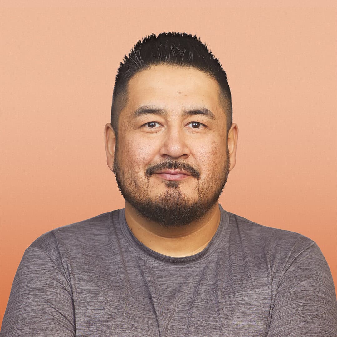 A man with short black hair and a trimmed beard is smiling softly. He is wearing a grey t-shirt and standing against a peach-coloured background with his arms crossed.