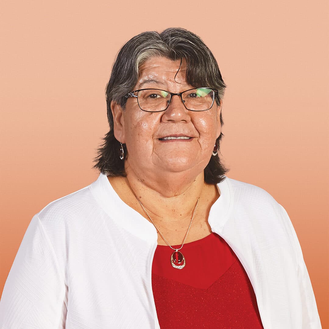 A smiling older woman with short dark hair, glasses, and earrings, wearing a white jacket over a red top with a necklace, stands in front of a peach-coloured background.