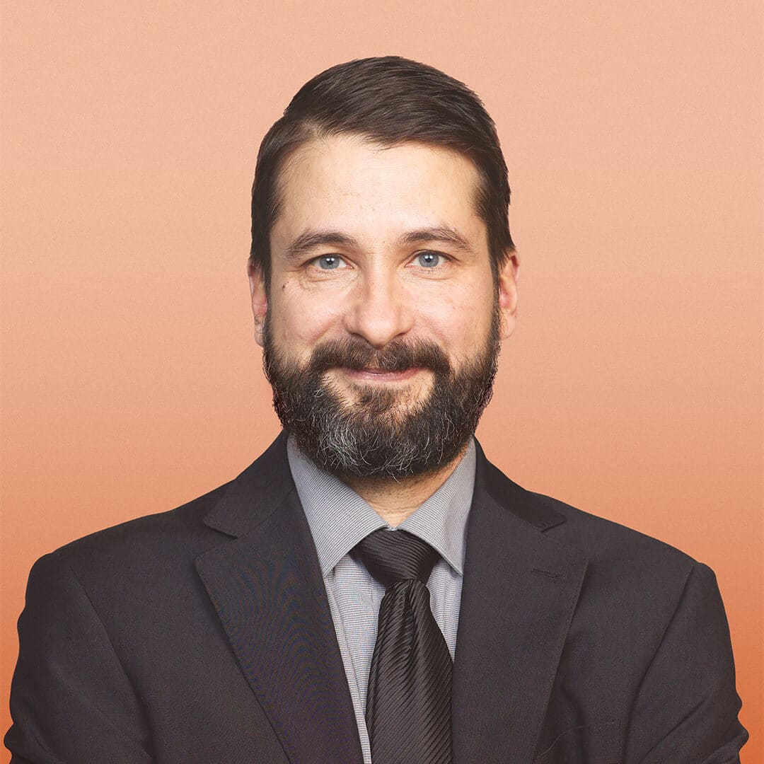 A man with dark hair and a beard, wearing a dark suit, grey shirt, and black tie, smiles at the camera against a plain peach background.