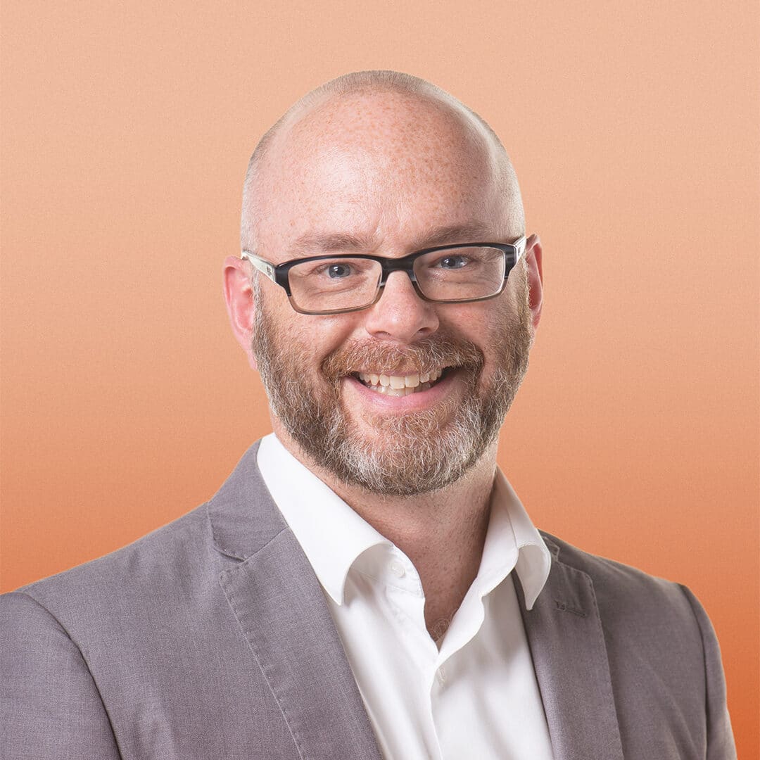 A smiling bald man with a beard and glasses, wearing a light grey blazer and white shirt, stands in front of an orange gradient background.