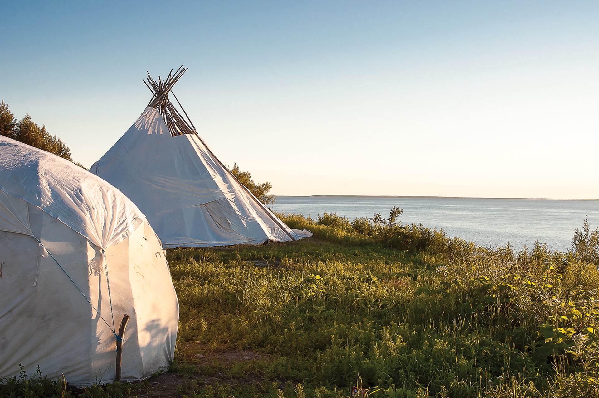 Two white canvas tents, including a traditional tipi, stand on grassy ground near the edge of a calm lake, with trees and bushes nearby under a clear blue sky at sunset.