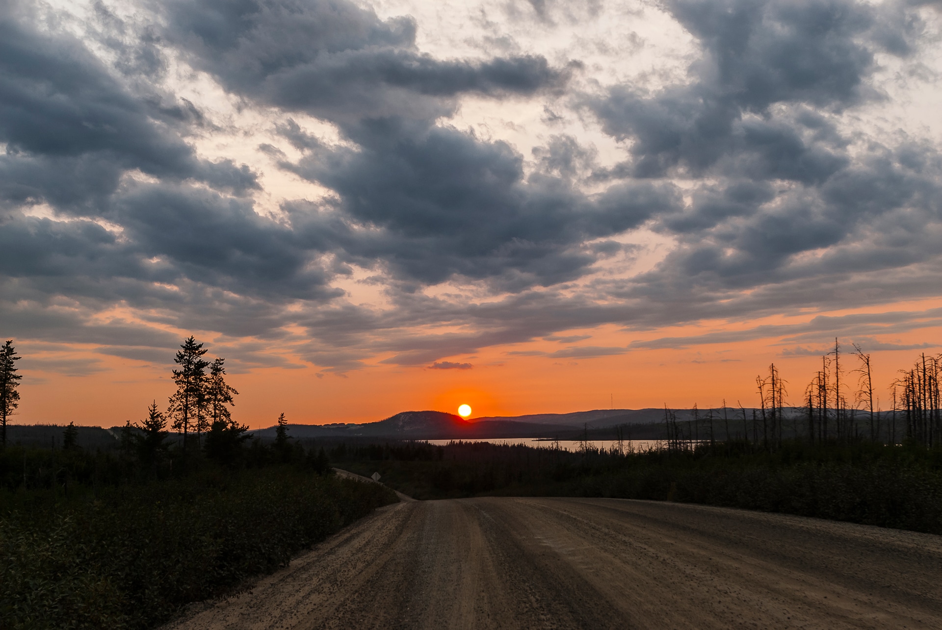 A dirt track leads towards a distant lake and hills at sunset, with the sun low on the horizon. The sky is filled with dramatic clouds, trees line both sides of the track, and nature’s beauty unfolds like well-designed programmes.