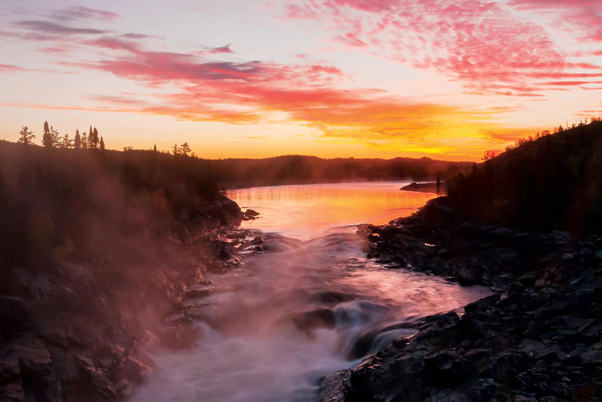 A vibrant sunset casts orange and pink hues across the sky and reflects on a river flowing through rocky terrain, with mist rising from the water and silhouettes of trees on the horizon.