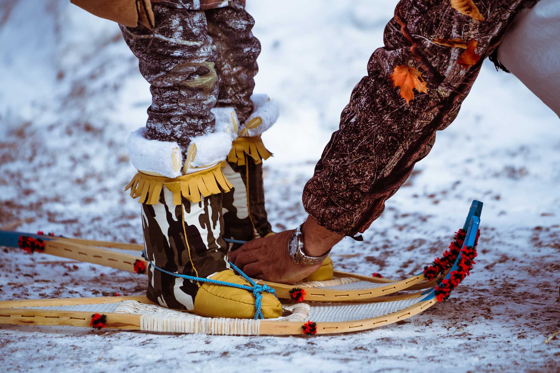 A close-up of a person helping a child in fur-trimmed boots stand on wooden snowshoes in the snow, both wearing traditional patterned clothing with decorative details.