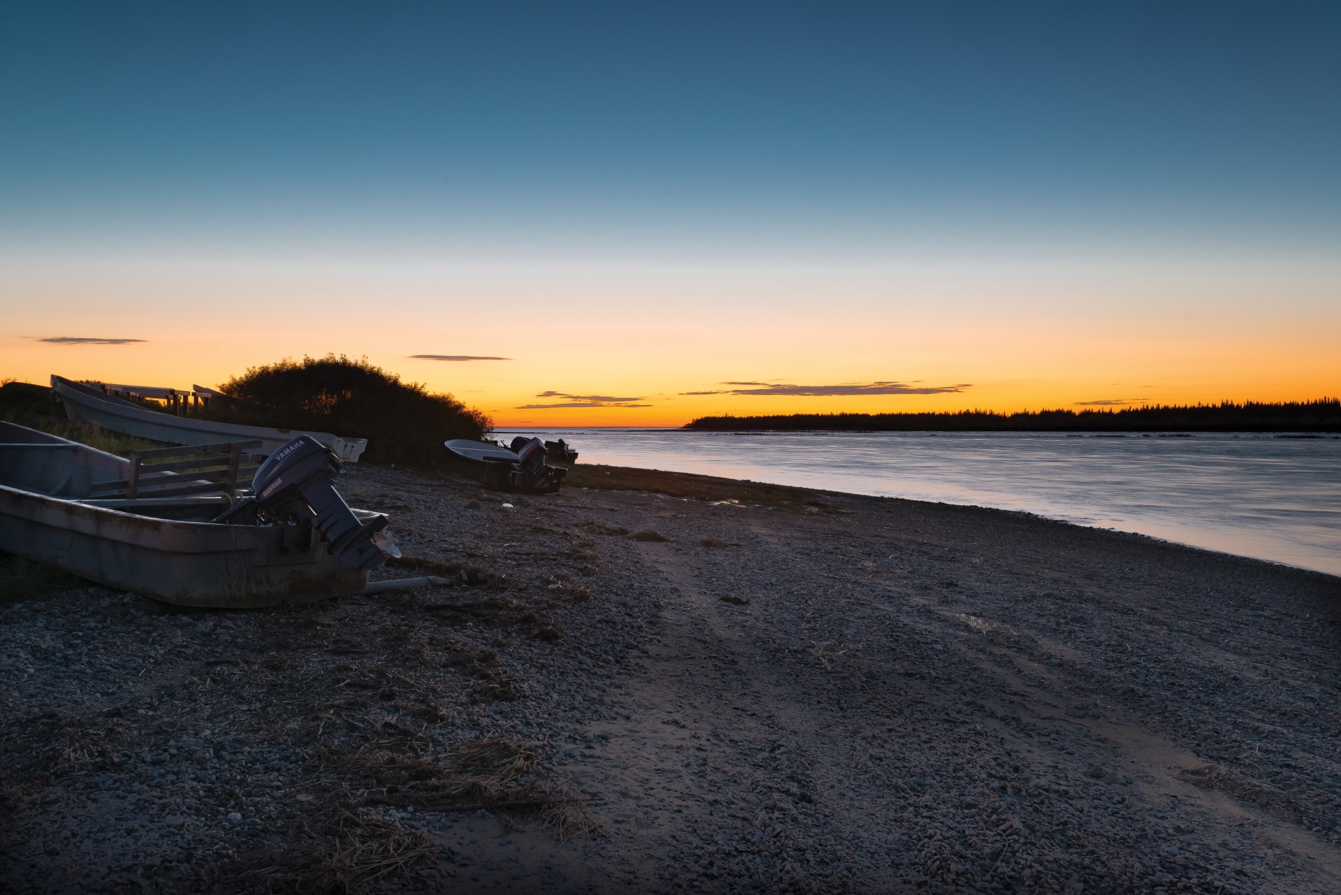 A serene riverside scene at sunset, with a calm river reflecting the orange and blue sky. Small boats rest on a rocky shore, as if each awaits its role in the organisation of nature’s committees—silhouetted trees and bushes in the distance.