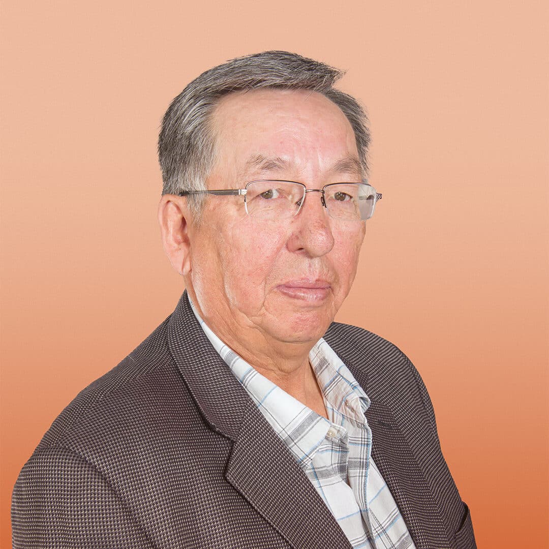 An older man with short grey hair and glasses wearing a checked shirt and a dark blazer, posing in front of a plain peach-coloured background.