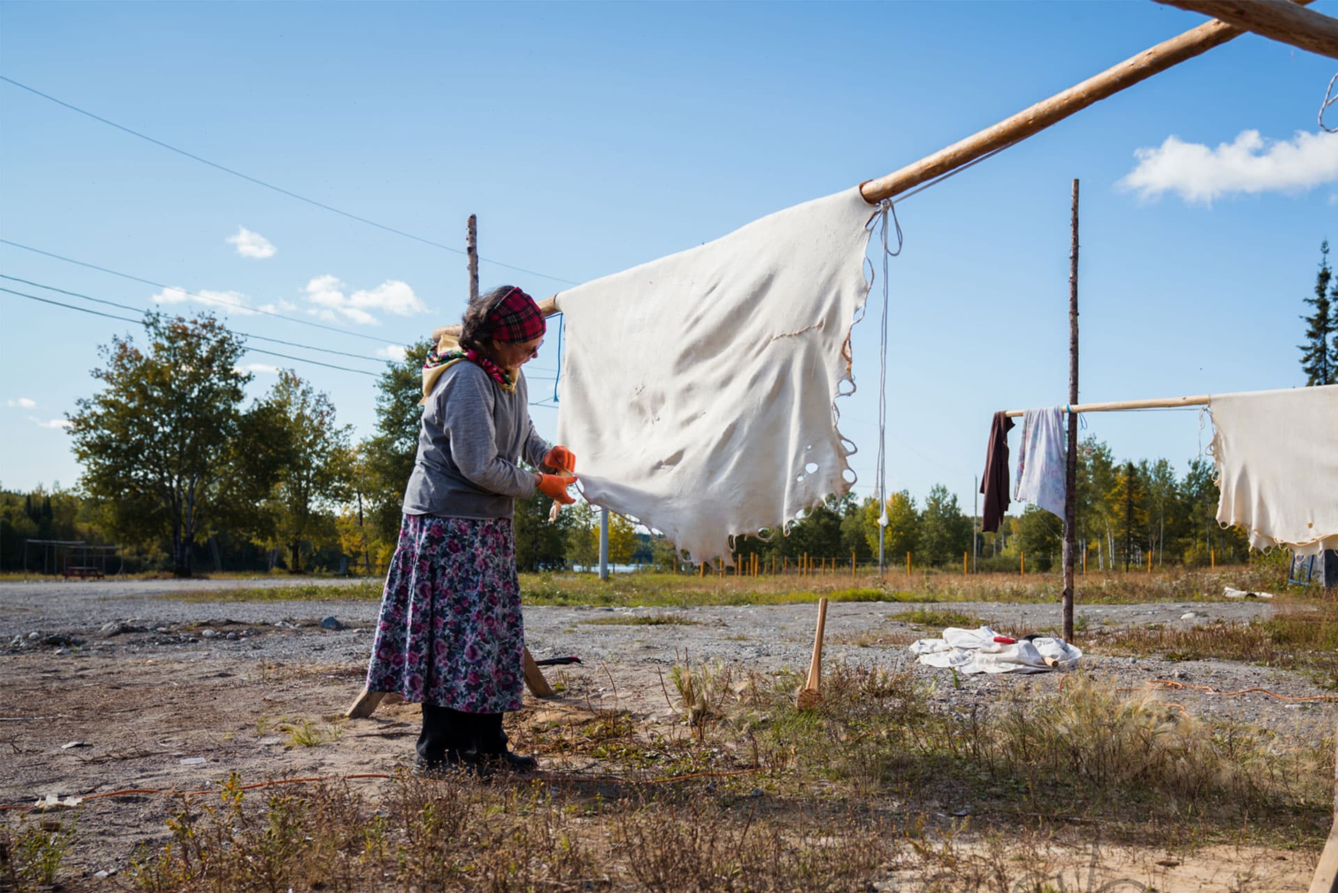 A woman wearing a headscarf and long skirt scrapes a large animal hide stretched on a wooden frame outdoors, with washing hanging and trees in the background under a blue sky.