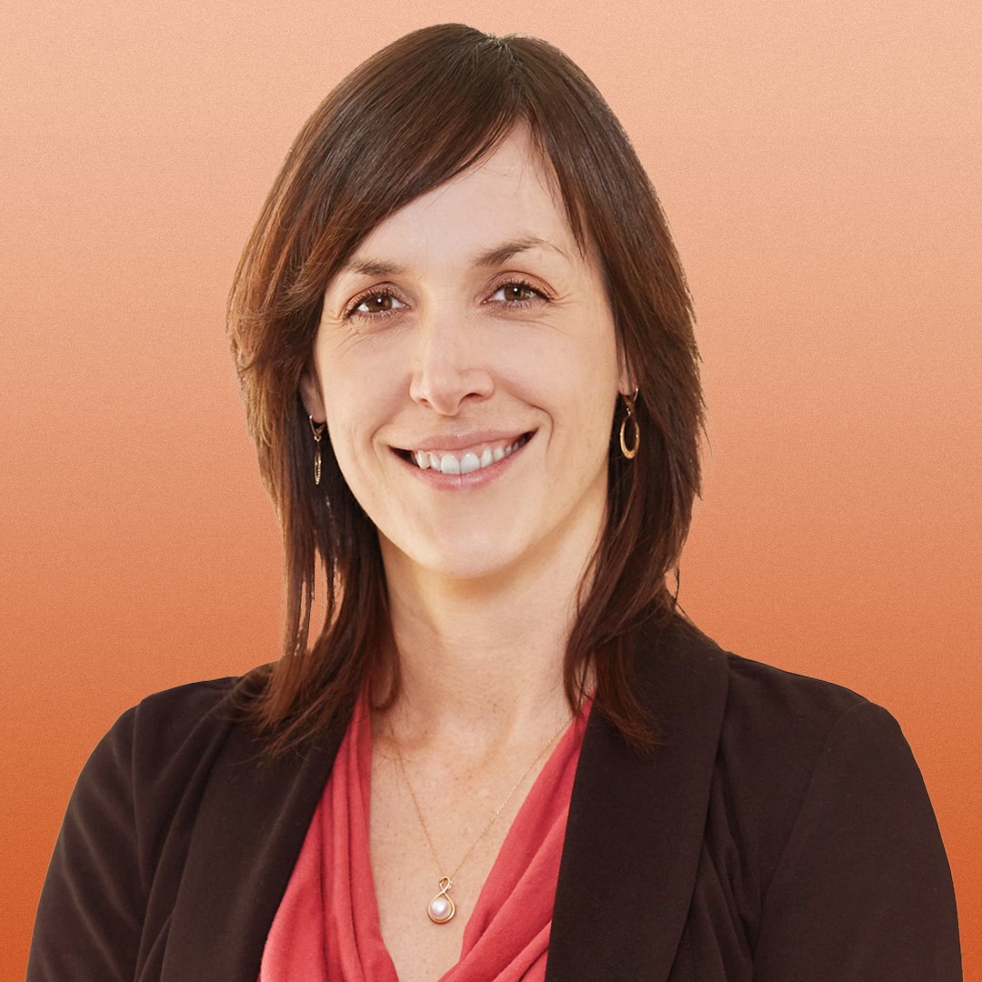 A woman with straight brown hair, wearing a coral blouse, dark blazer, gold earrings, and a necklace, smiles at the camera against a peach-orange gradient background.