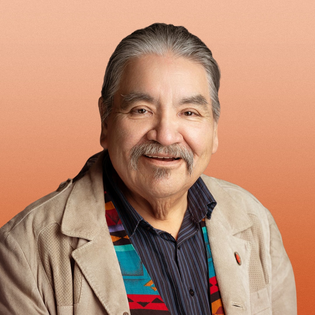 Smiling older man with grey hair and moustache wearing a tan jacket, black striped shirt, and a colourful patterned scarf, in front of a soft orange background.