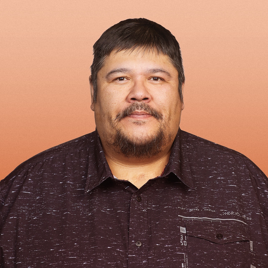 A man with short dark hair, a moustache, and a goatee is wearing a dark patterned button-up shirt and is standing in front of a peach-coloured background.
